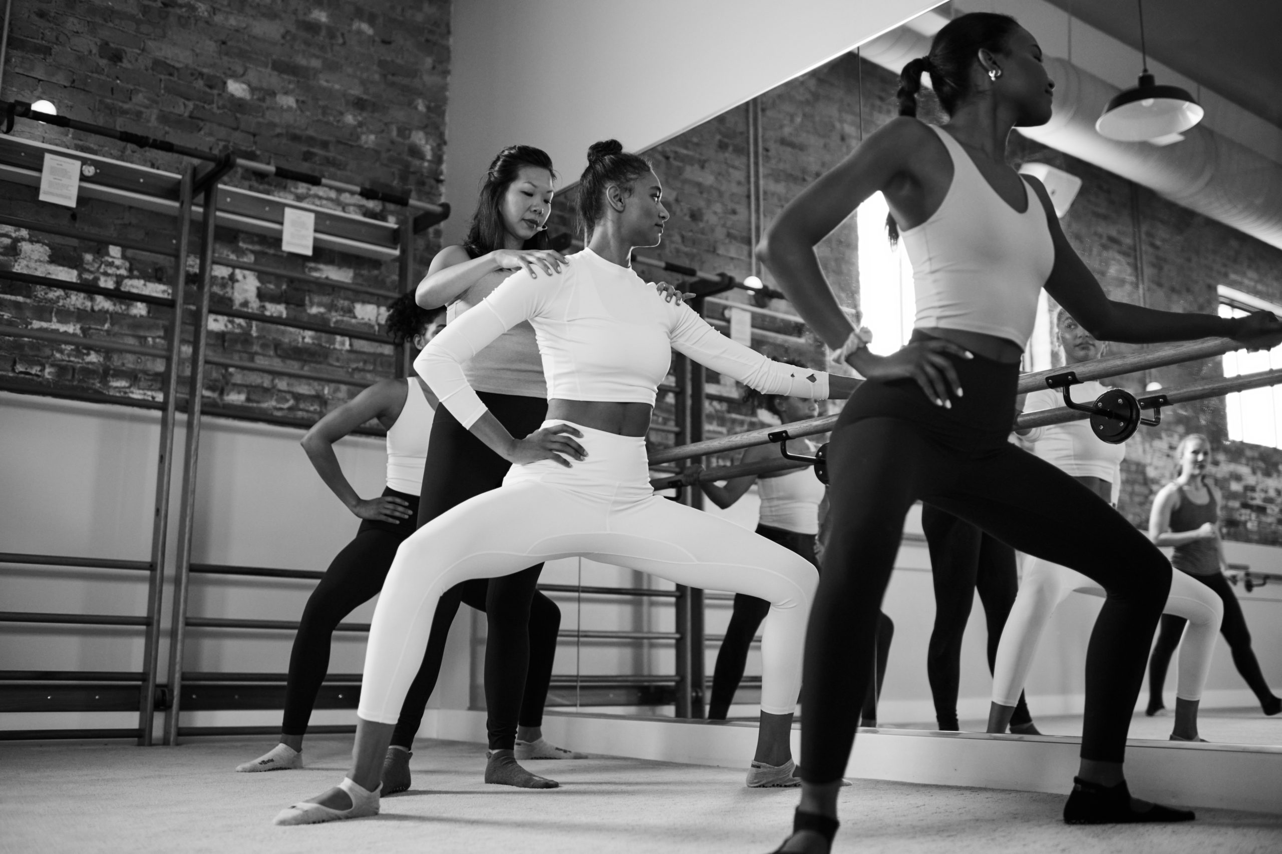 Black and white image of instructor correcting a members form at the barre in a Bar Method studio